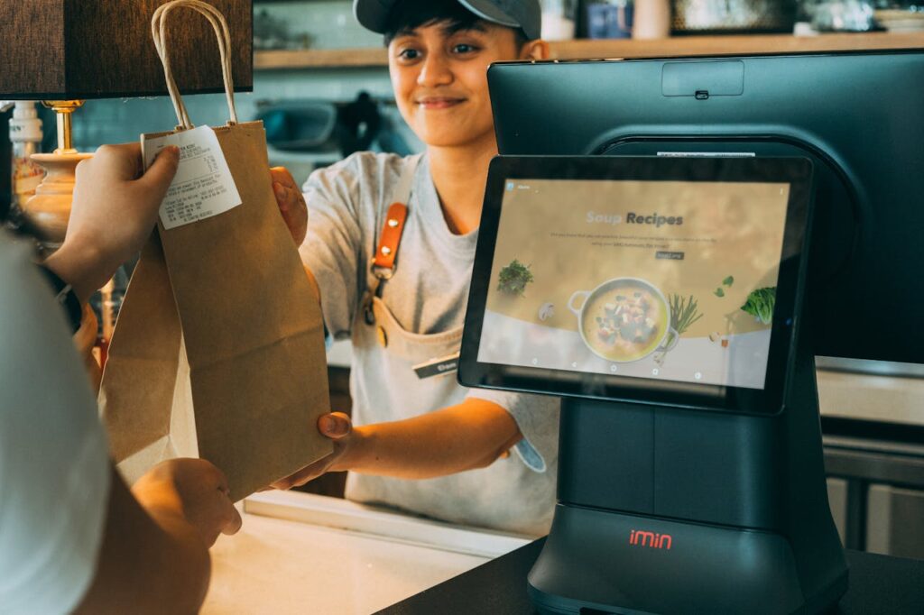 Smiling café employee handing a takeout bag to a customer across the counter, representing how between-song messaging supports better in-store experiences and sales.