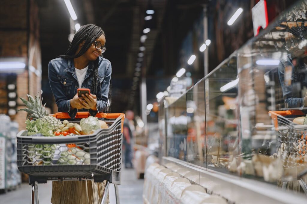 Customer shopping in a grocery store aisle using a smartphone and cart filled with fresh produce
