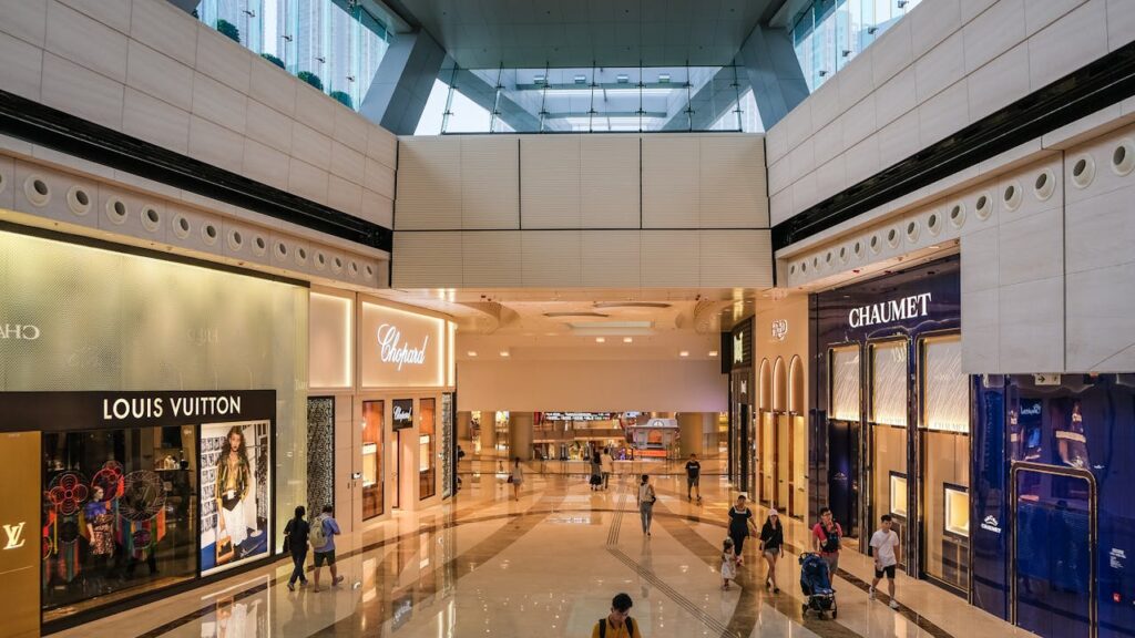 Modern shopping mall interior with luxury retail stores and shoppers walking through polished marble floors.